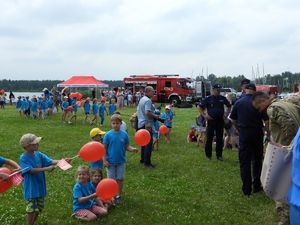 Trawiasty plac na który widać dużą grupę dzieci z czerwonymi balonami i opiekunkami, Kilkoro Policjantów w mundurach. W tle czerwony namiot oraz różne samochody w tym wóz strażacki.