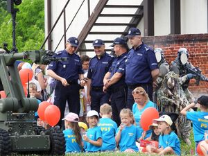 Dzieci w wieku przedszkolnym wraz z opiekunką ubrane w niebieskie koszulki z napisem Wieliszew PS1, siedzące na trawie z czerwonymi balonami w dłoni. Za nimi stoją umundurowani policjanci - Zastępca Komendanta Głównego Policji, Dyrektor Biura Prewencji KGP, Zastępca Komendanta Stołecznego Policji, Komendant Komisariatu Rzecznego Policji w Warszawie. Na pierwszym planie stoi robot rozpoznania saperskiego.
