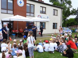 Kilkoro policjantów stojących przed grupą dzieci siedzących na trawie. Kilkoro zgłasza się do odpowiedzi podnosząc rękę ku górze. W tle ratownicy WOPR w czerwonych koszulkach.