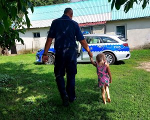 dziecko idące za rękę z policjantem, w tle budynek mieszkalny i radiowóz