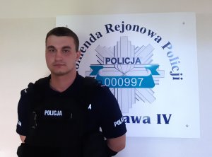 sierż. Maciej Krassowski stoi na tle ściany z logiem Komendy Rejonowej Policji Warszawa IV