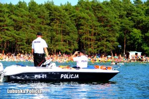 policjanci na policyjnej łodzi pływają przy brzegu, w tle grupa młodzieży i dzieci