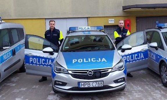 trzy radiowozy policyjne , obok jednego z nich stoja dwaj policjanci
