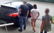 policjant i policjantka prowadzą do samochodu  zatrzymaną kobietę, która ma nałożone kajdanki na ręce trzymane z tyłu