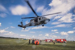 Strażacy napełniają zbiornik bambi bucket wężami, nad nimi unosi się śmigłowiec.