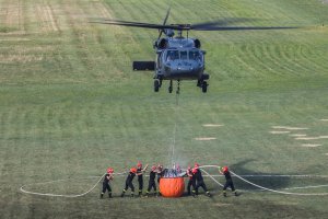 Strażacy napełniają bambi bucket wężami, nad nimi unosi się śmigłowiec widziany z przodu.