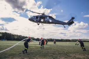 Strażacy na ramionach podciągają węże do napełnienie bambi bucket, w drugim planie śmigłowiec z opuszczonym zbiornikiem na wodę na płytę lądowiska.