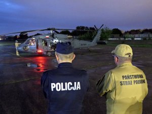 Zastępca Komendanta Głównego Policji nadinsp. Romana Kustera oraz zastępca Komendanta Głównego Państwowej Straży Pożarnej nadbryg. Krzysztof Hejduk obserwują przygotowania do nocnego startu śmigłowca S70i Black Hawk, widocznego na drugim planie.