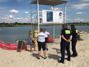 Policjanci wraz ze strażnikami miejskimi stoją przy wieżyczce ratowników wodnych i obserwują akwen wodny
