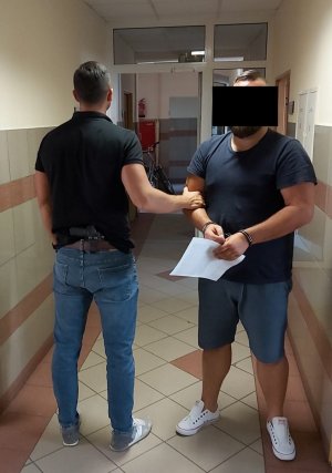 policjant prowadzi zatrzymanego mężczyznę - policjant stoi tyłem, mężczyzna jest odwrócony przodem, na rękach ma kajdanki a w dłoniach trzyma kartkę