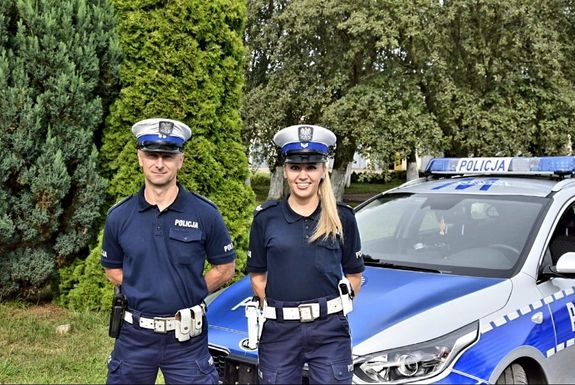 Umundurowana policjanta z policjantem z ruchu drogowego stoją obok oznakowanego radiowozu.