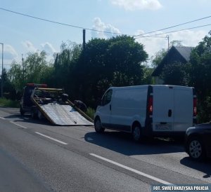 Samochód dostawczy koloru białego stoi obok samochodu typu laweta przygotowujący się do wjazdu na lawetę.