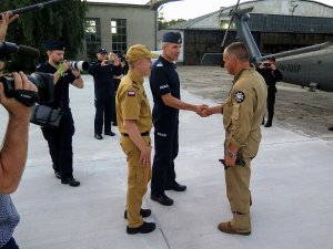 zastępca komendanta głownego policji wita policyjnych lotników, w tle inni policjanci wykonujący zdjęcia, po prawej stronie policyjny Black Hawk