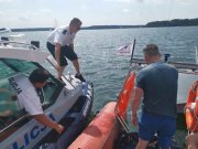 Umundurowany policjant w pozycji pochylonej, przygotowujący się do skoku na łódź wodująca obok motorówki z napisem Policja.