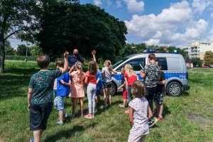 policjant z grupą dzieci stoi przy radiowozie podczas spotkania plenerowego i rozmawia o bezpieczeństwie