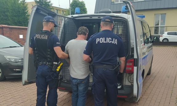 Zdjęcie przedstawia dwóch umundurowanych policjantów prowadzących zatrzymanego mężczyznę do więźniarki