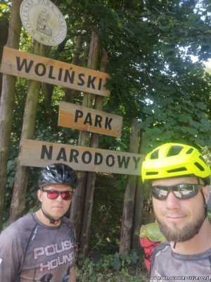 Dwaj mężczyźni w kaskach rowerowych przy napisie umieszczonym na drewnianych drogowskazach: Woliński Park Narodowy