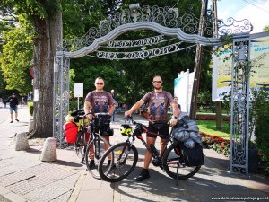 Dwaj mężczyźni w kaskach z rowerami przy ozdobnej bramie z napisem: Międzyzdroje, Park Zdrojowy