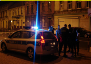 radiowóz policyjny z włączonymi sygnałami stoi obok policjantów i innych osób obok budynku