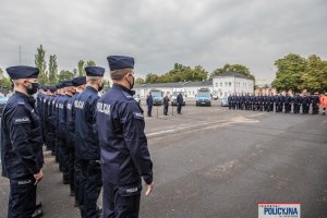 policjanci zebrani na placu stoją w dwuszeregu, przy mikrofonach Minister SWiA i Komendant Główny Policji, z tylu widoczne radiowozy