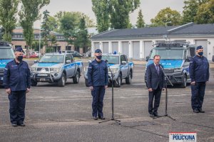 przy mikrofonach Minister SWiA i Komendant Główny Policji, z boku dwaj umundurowani policjanci, z tyłu widoczne radiowozy