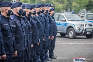 policjanci zebrani na placu stoją w dwuszeregu, z tyłu widoczne radiowozy
