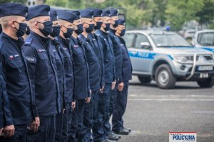 policjanci zebrani na placu stoją w dwuszeregu, z tyłu widoczne radiowozy