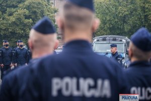 policjanci zebrani na placu stoją w dwuszeregu - widok z tyłu, przed nimi radiowozy