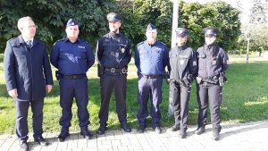 Pięcioosobowa grupa umundurowanych policjantów i policjantek polskich oraz czeskich na tle zieleni parku, którym towarzyszy mężczyzna w eleganckim płaszczu.