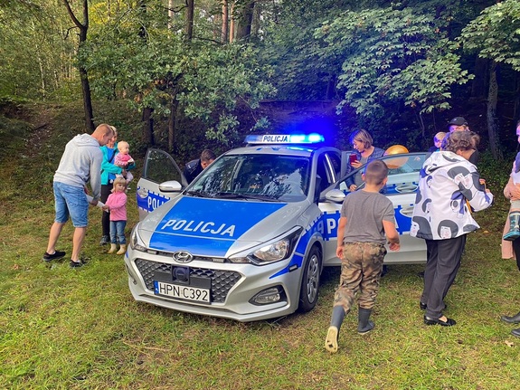 policjanci rozmawiają o bezpieczeństwie podczas spotkania profilaktycznego, dzieci i młodzież oraz osoby dorosłe oglądają policyjny radiowóz