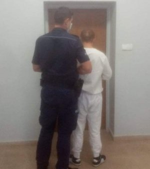 Na korytarzy aresztu przed drzwiami  pokoju przesłuchań stoi mężczyzna w białej buzie i spodniach za nim umundurowany policjant