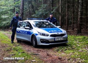 Na zdjęciu umundurowani policjanci stojący przy oznakowanym radiowozie w lesie.