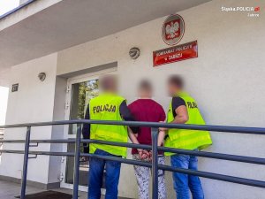 Nieumundurowani policjanci, w kamizelkach odblaskowych z napisem policja, prowadzą zatrzymanego mężczyznę.