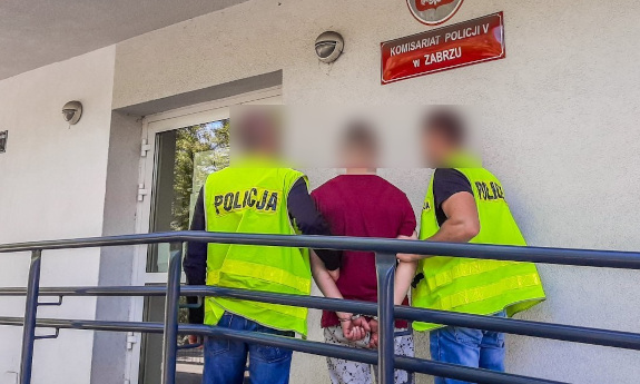 Nieumundurowani policjanci, w kamizelkach odblaskowych z napisem policja, prowadzą zatrzymanego mężczyznę.