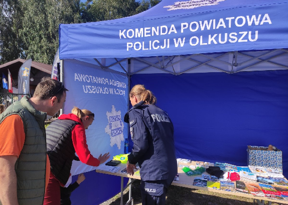 Policjantka przy stanowisku profilaktycznym pod niebieskim namiotem z napisem Komenda Powiatowa Policji w Olkuszu rozmawia z osobami o bezpieczeństwie pokazując broszury. Kobieta wykonuje odbitkę linii papilarnych.