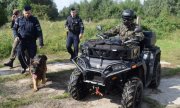 żołnierze Wojsk Obrony Terytorialnej i policjanci podczas ćwiczeń w miejscowości Trzebiatów na terenie wojskowym. Jeden z funkcjonariuszy wojska jedzie na quadzie, drugi wraz z dwoma umundurowanymi policjantami idzie obok. Jeden z policjantów prowadzi na smyczy psa