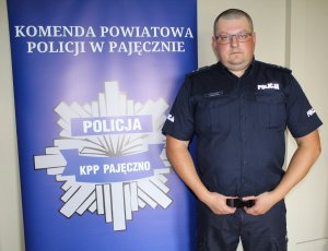 Umundurowany policjant stoi na tle baneru z napisem Komenda Powiatowa Policji w Pajęcznie