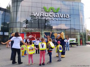 Pod centrum handlowym dzieci wspólnie z komisarzem lwem i policjantkami i animatorem tańczą i śpiewają.