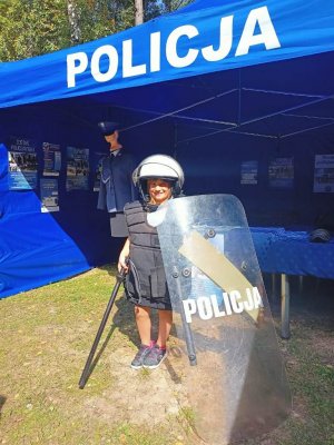 Namiot z napisem Policja. W środku stoi dziecko ubrane w policyjną kamizelkę taktyczną, hełm policyjny, w lewym ręku trzyma policyjną tarczę, w prawym pałkę. Z tyłu widoczny jest  manekin ubrany  w policyjny mundur, a na ścianach namiotu przypięte są ulotki