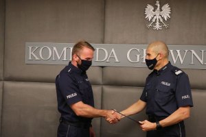 Zastępca Komendanta Głównego Policji w mundurze polowym ściska dłoń umundurowanemu policjantowi i wręcza mu teczkę. Z tyłu na ścianie widoczny fragment napisu: Komenda Główna Policji, a nad nim orzeł w koronie