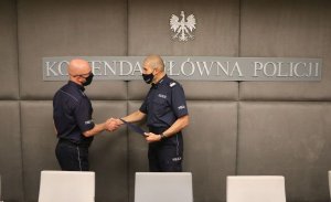 Zastępca Komendanta Głównego Policji w mundurze polowym ściska dłoń umundurowanemu policjantowi i wręcza mu teczkę. Z tyłu na ścianie widoczny fragment napisu: Komenda Główna Policji, a nad nim orzeł w koronie