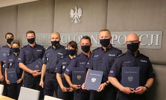 Zastępca Komendanta Głównego Policji w mundurze i siedmioro umundurowanych policjantów, w tym dwie kobiety, stoją jeden za drugim pod ścianą z widocznymi fragmentami napisu: Komenda Główna Policji, a nad nim orzeł w koronie. Dziesięcioro policjantów w dłoniach trzyma granatową teczkę