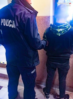 umundurowany policjant trzyma za ramię zakutego w kajdanki mężczyznę podejrzanego o usiłowania zabójstwa