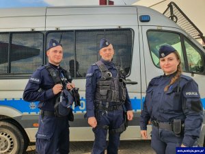 Na pierwszym planie troje funkcjonariuszy policji w mundurach, za nimi stojący radiowóz