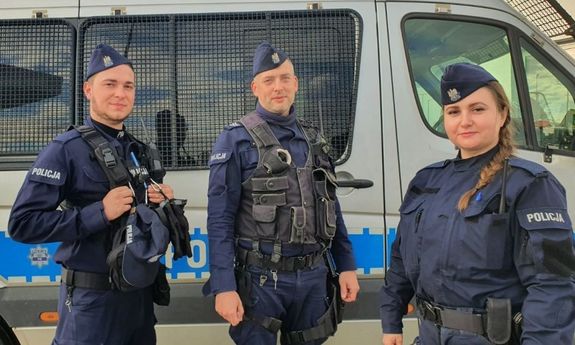 Na pierwszym planie troje funkcjonariuszy policji w mundurach, za nimi stojący radiowóz