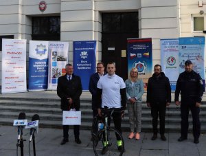 Mł. asp. Sebastian Nowacki, funkcjonariusz Komendy Miejskiej Policji w Płocku, stojący z rowerem. Za nim stoją policjanci i samorządowcy uczestniczący w rozpoczęciu trasy rowerowej, w celu zebrania pieniędzy na leczenie nastolatki.