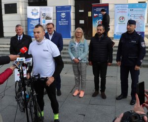 Mł. asp. Sebastian Nowacki, funkcjonariusz Komendy Miejskiej Policji w Płocku, stojący z rowerem. W tle policjanci i samorządowcy uczestniczący w rozpoczęciu trasy rowerowej, w celu zebrania pieniędzy na leczenie nastolatki.
