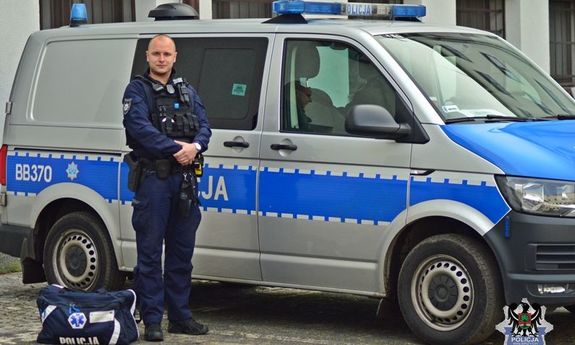 Umundurowany policjant stojący przy radiowozie, obok niego torba medyczna