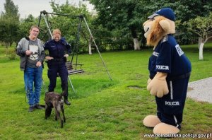 Na podwórku policjantka wraz z psem tropiącym którego trzyma na smyczy chłopiec, obok maskotka policyjna