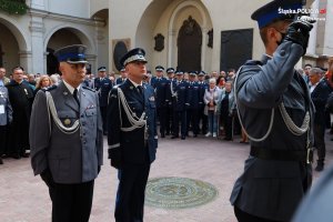 Stojący na baczność trzej policjanci, dookoła nich ludzie, ksiądz, grupa funkcjonariuszy zebrana na placu